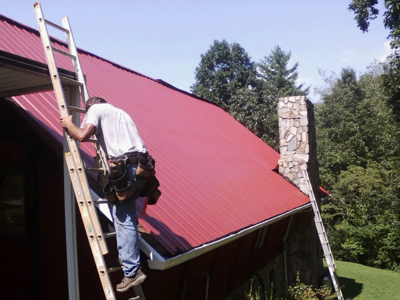 Roofer on a ladder beside a red metal roof and stone chimney — Soffit & Fascia Repair by Roofing Brothers in Pinopolis